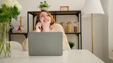 Genç kızıl saçlı kadın dizüstü bilgisayar kullanıyor ve evdeki akıllı telefondan konuşuyor.