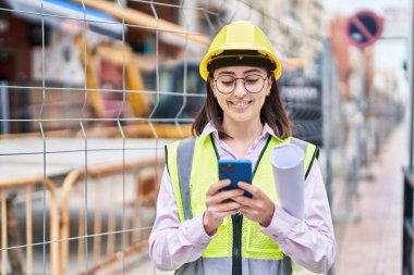 Young hispanic woman architect using smartphone holding blueprints at street