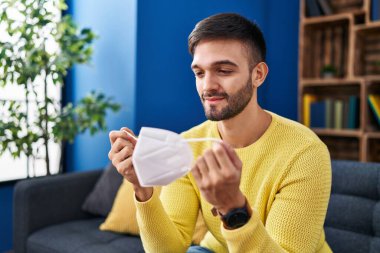 Evdeki kanepede oturan tıbbi maske takan İspanyol genç adam.