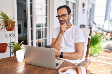 Orta yaşlı bir adam bilgisayarı evde kullanıyor kameraya kendinden emin bakıyor kollarını kavuşturup gülümsüyor ve çenesini kaldırıyor. Olumlu düşünmek. 