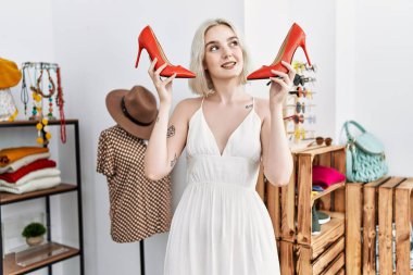 Giysi mağazasında yüksek topuklu ayakkabı tutan beyaz bir kadın gülümsüyor.