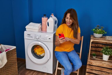 Çamaşır odasında çamaşır makinesi bekleyen akıllı telefon kullanan genç Latin kadın.