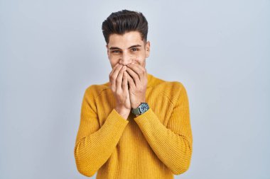 Mavi arka planda dikilen Latin asıllı genç adam gülüyor ve utanıyor kıkırdıyor elleriyle ağzını kapatıyor, dedikodu ve skandal konsepti yapıyor. 