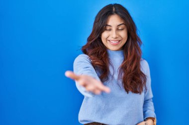 Hispanic young woman standing over blue background smiling cheerful offering palm hand giving assistance and acceptance. 