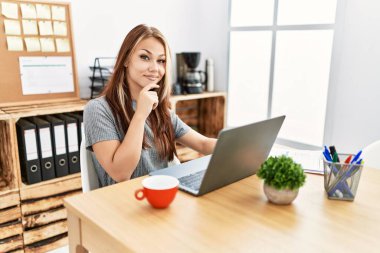 Ofiste laptopuyla çalışan genç esmer kadın kameraya kollarını kavuşturup gülümseyen ve çenesini kaldıran kendinden emin bir şekilde bakıyor. Olumlu düşünmek. 