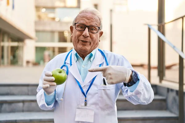 老年医生 白头发 拿着健康的绿色苹果 手指对着一个笑得开心而自豪的人 — 图库照片