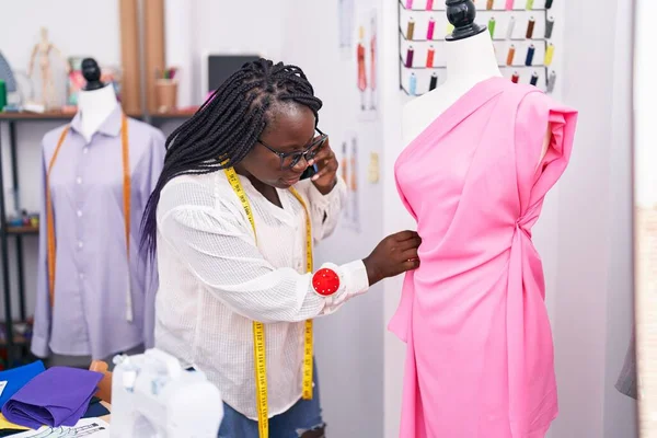 African american woman tailor talking on smartphone sewing dress at tailor shop