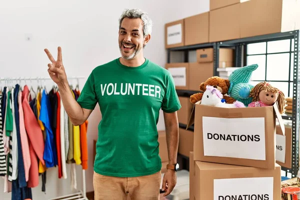 Middle age hispanic man wearing volunteer t shirt at donations stand smiling with happy face winking at the camera doing victory sign with fingers. number two.