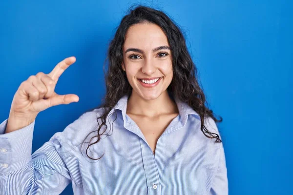 若いブルネットの女性は青い背景の笑顔の上に立っており 指を見てカメラで小さなサイズのサインをする手で自信を持ってジェスチャー 概念を測定し — ストック写真