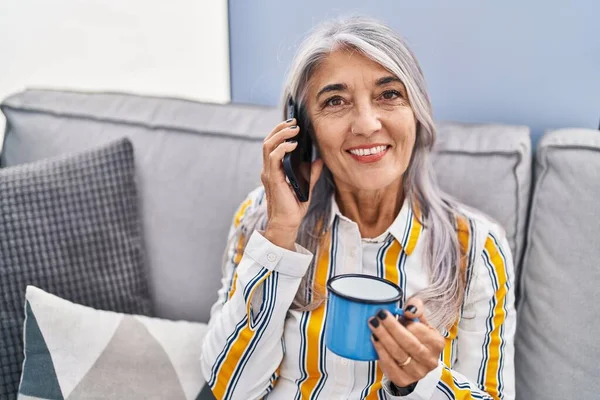 Middle age grey-haired woman drinking coffee and talking on the smartphone at home