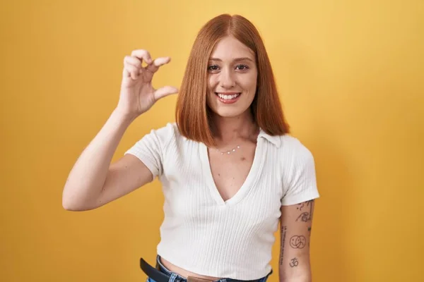 年轻的红头发女人站在黄色的背景上 微笑着 自信地做手势 手拿着小尺寸的标志 手拿着手指 手拿着相机 计量概念 — 图库照片