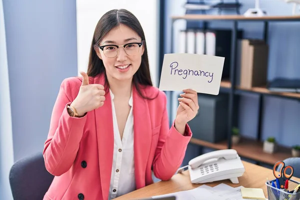 Chinese young woman working at the office holding pregnancy sign smiling happy and positive, thumb up doing excellent and approval sign