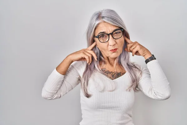 Middle age woman with grey hair standing over white background covering ears with fingers with annoyed expression for the noise of loud music. deaf concept.