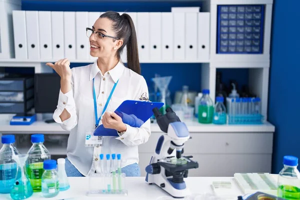 Young brunette woman working at scientist laboratory smiling with happy face looking and pointing to the side with thumb up.