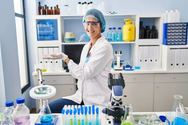 Brunette woman working at scientist laboratory pointing aside with hands open palms showing copy space, presenting advertisement smiling excited happy