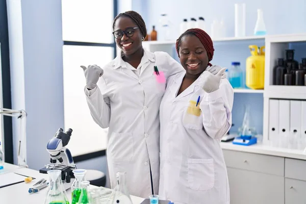 Two african women working at scientist laboratory pointing thumb up to the side smiling happy with open mouth