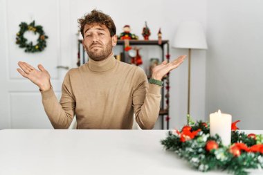 Noel süslemesi masasında oturan sakallı, genç yakışıklı ve elleri ve kolları havada, kafası karışmış bir adam. şüphe kavramı. 