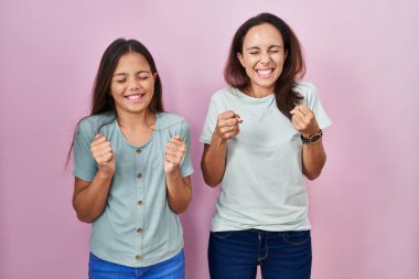 Young mother and daughter standing over pink background excited for success with arms raised and eyes closed celebrating victory smiling. winner concept. 