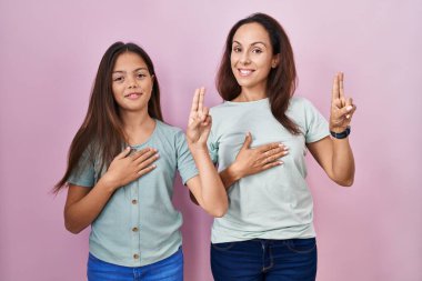 Young mother and daughter standing over pink background smiling swearing with hand on chest and fingers up, making a loyalty promise oath 