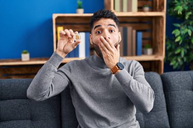 Hispanic man with beard holding virtual currency bitcoin covering mouth with hand, shocked and afraid for mistake. surprised expression 