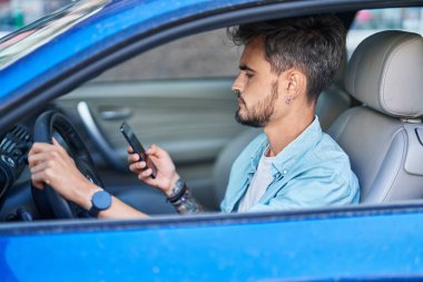 Young hispanic man using smartphone sitting on car at street