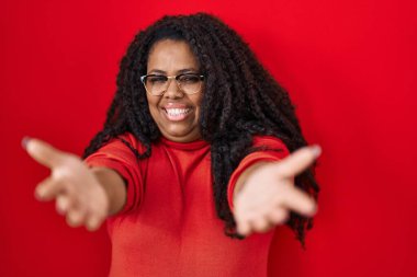 Plus size hispanic woman standing over red background looking at the camera smiling with open arms for hug. cheerful expression embracing happiness. 