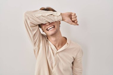 Young man standing over isolated background covering eyes with arm smiling cheerful and funny. blind concept. 