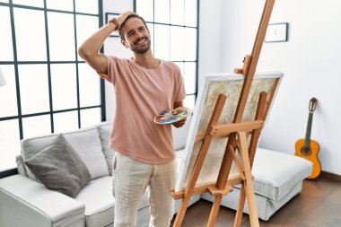 Young hispanic man with beard painting on canvas at home smiling confident touching hair with hand up gesture, posing attractive and fashionable 