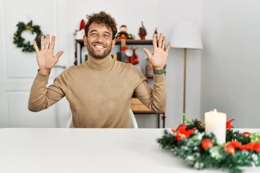 Noel süslemesi masasında oturan sakallı yakışıklı genç adam on numarayı gösterip işaret ederken kendinden emin ve mutlu bir şekilde gülümsüyor.. 