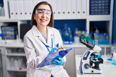 Middle age woman scientist smiling confident writing on clipboard at laboratory