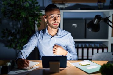 Young hispanic man working at the office at night smiling looking to the side and staring away thinking. 