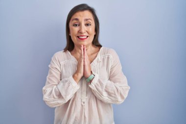 Orta yaşlı Latin bir kadın mavi arka planda duruyor. Birlikte dua ediyor, af diliyor. Kendine güvenen bir gülümseme.. 