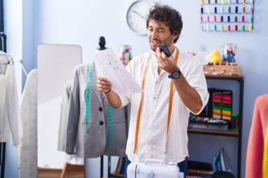 Genç İspanyol terzi akıllı telefondan konuşuyor giyim fabrikasında kıyafet tasarımı yapıyor.