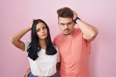 Young hispanic couple standing over pink background confuse and wondering about question. uncertain with doubt, thinking with hand on head. pensive concept. 