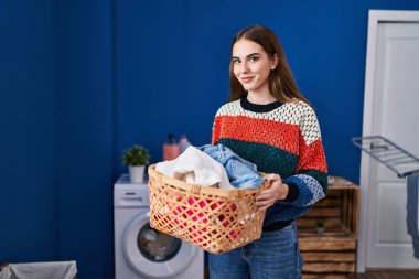 Çamaşır odasında çamaşır sepetini tutan kendine güvenen genç bir kadın.