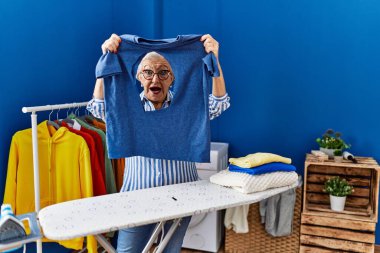 Çamaşırhanede yakılmış demir t-shirt tutan yaşlı, gri saçlı kadın.