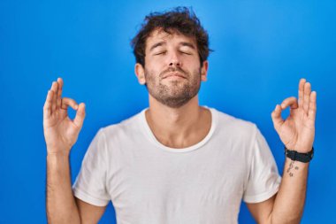 Mavi arka planda duran İspanyol genç adam rahatlamış ve parmaklarıyla meditasyon hareketi yaparken gözleri kapalı gülümsüyordu. yoga konsepti. 