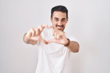Handsome hispanic man standing over white background smiling in love doing heart symbol shape with hands. romantic concept. 