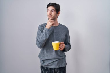Young hispanic man wearing pajama drinking a cup of coffee with hand on chin thinking about question, pensive expression. smiling with thoughtful face. doubt concept. 