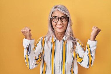 Sarı arka planda duran gri saçlı orta yaşlı bir kadın. Gözlüklü, şaşırmış ve şaşırmış bir şekilde kolları açık, gözleri açık bir şekilde başarıyı kutluyor. Kazanan konsept. 