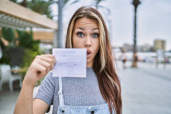 Young caucasian woman holding covid record card scared and amazed with open mouth for surprise, disbelief face