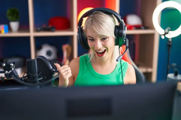 若いですCaucasian女性再生ビデオゲームを身に着けていますヘッドフォンを指差す親指へザ側笑顔幸せとともにオープン口 — ストック写真