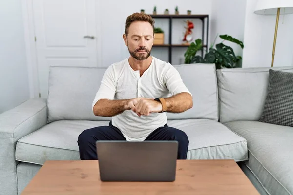 Middle age man using laptop at home checking the time on wrist watch, relaxed and confident