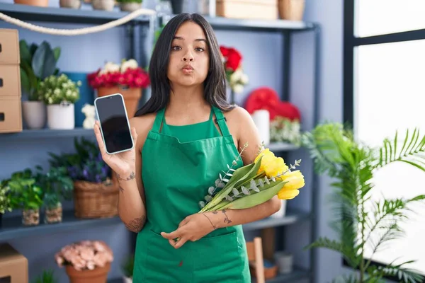 Brunette woman working at florist shop holding smartphone puffing cheeks with funny face. mouth inflated with air, catching air.
