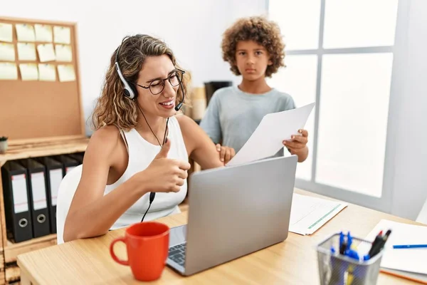 Young business mother working at the office with kid smiling happy and positive, thumb up doing excellent and approval sign