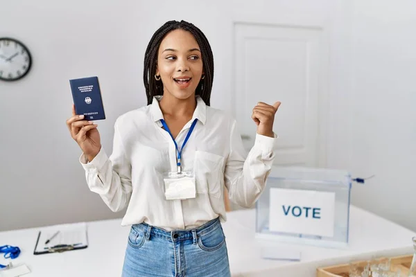 Young african american woman at political campaign election holding deutschland passport pointing thumb up to the side smiling happy with open mouth