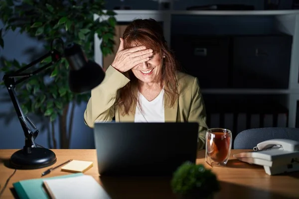 Middle age hispanic woman working using computer laptop at night smiling and laughing with hand on face covering eyes for surprise. blind concept.