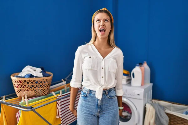 Young blonde woman at laundry room angry and mad screaming frustrated and furious, shouting with anger. rage and aggressive concept.