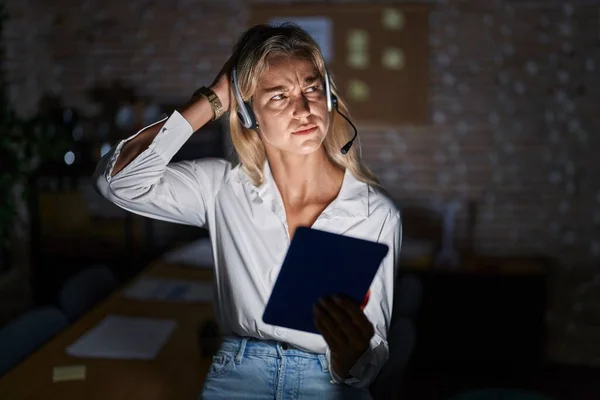 Young blonde woman working at the office at night confuse and wondering about question. uncertain with doubt, thinking with hand on head. pensive concept.