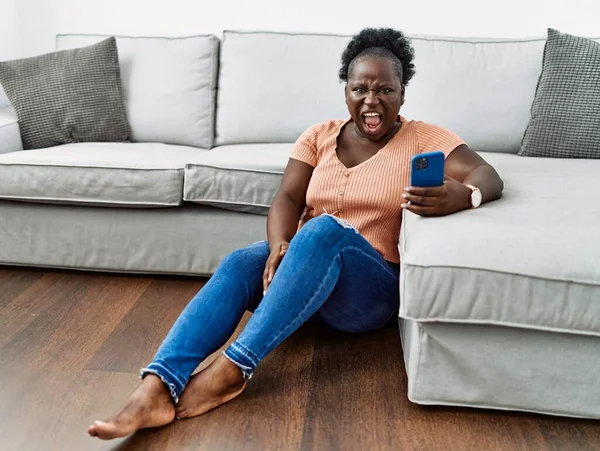 Young african woman using smartphone sitting on the floor at home angry and mad screaming frustrated and furious, shouting with anger. rage and aggressive concept.